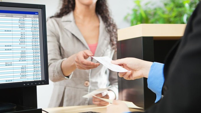Over-the-shoulder shot of a transaction between a bank teller and a customer in a retail bank. The teller is wearing a black suit and receiving a check from the tan-suited customer over the bank counter window. Photographed in horizontal format.