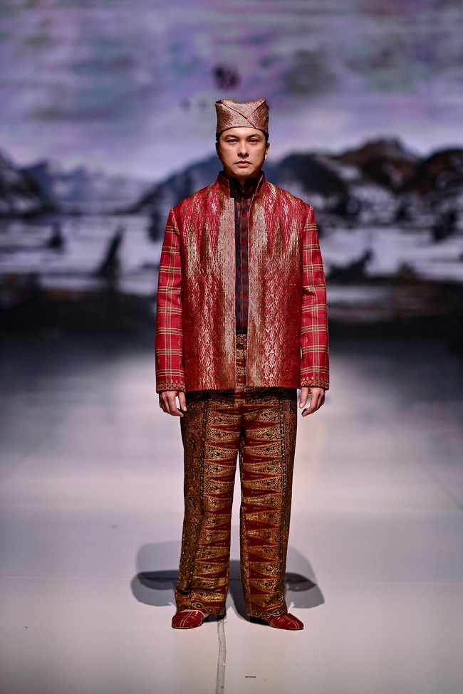 Peragaan karya Auguste Soesastro di panggung Dewi Fashion Knights 2025 ditutup dengan penampilan aktor Nicholas Saputra. Berlenggang di catwalk, ia memeragakan setelan merah dengan jas tak berkancing yang elegan, dipadu celana panjang songket yang terasa mewah berkat jahitan benang emas. (Foto: Dok. Jakarta Fashion Week)
