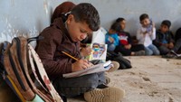 Siswa menulis di buku catatan dengan lutut sebagai meja di antara reruntuhan sekolah. Kekurangan sarana prasarana dan bahan ajar menjadi tantangan besar bagi pendidikan di Idlib. Foto: REUTERS/Karam al-Masri