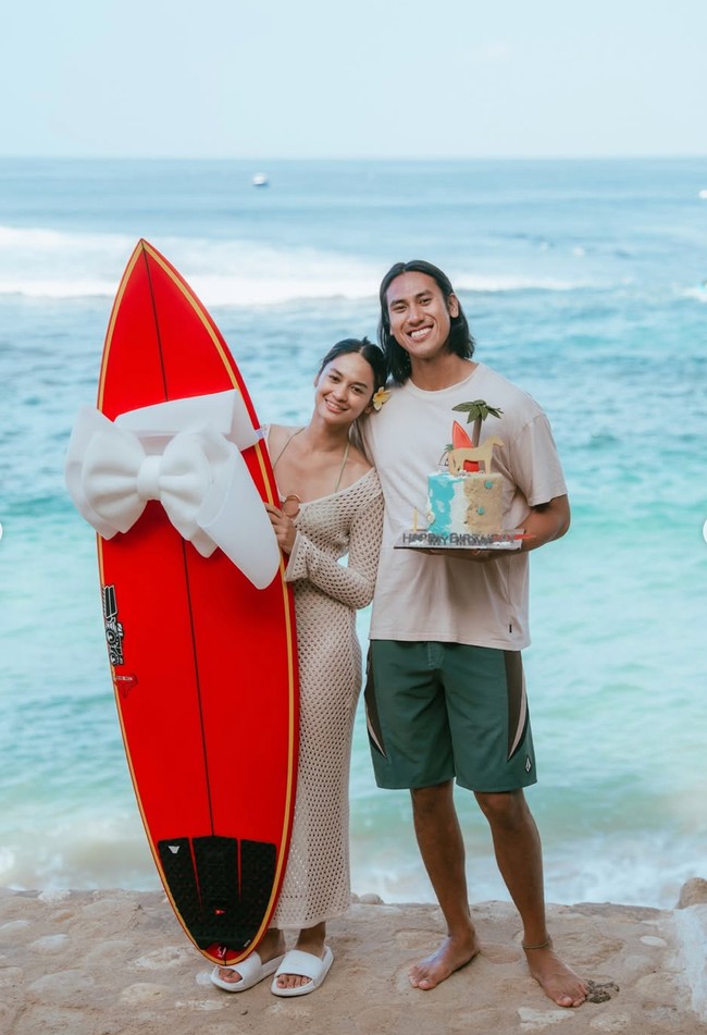 Kue ulang tahun dengan tema pantai terlihat dipegang oleh Sean. Sementara Hana memegang papan surfing merah yang sudah dihias dengan pita putih untuk diberikan kepada Sean sebagai kado ulang tahun. Foto: Instagram/@hanamalasan