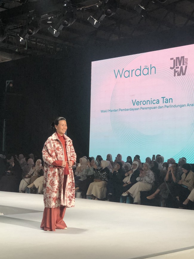 Veronica Tan, Wakil Menteri Pemberdayaan Perempuan dan Perlindungan Anak jadi model di JMFW 2026 mengenakan setelan celana wide-leg pants berwarna terracotta dengan atasan berwarna senada di dalamnya. Statement piece dari outfit ini adalah jaket atau coat panjang berwarna putih dengan motif bunga-bunga merah yang bold. Tampilannya cerah, playful, dan modis. Foto: Gresnia/Wolipop.