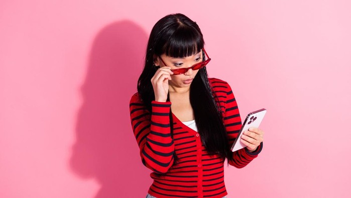 Stylish woman wearing a red striped sweater and holding a phone, standing against a bright pink background with a curious expression.
