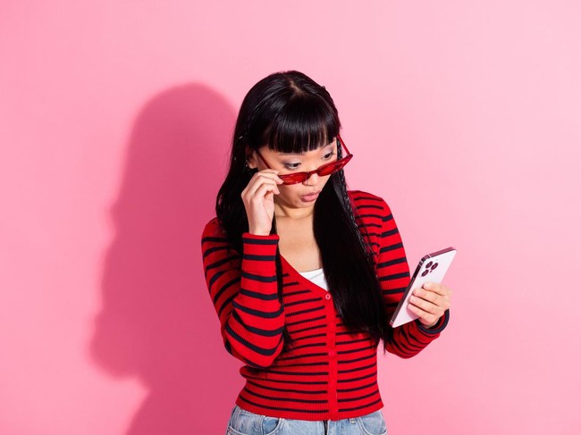 Stylish woman wearing a red striped sweater and holding a phone, standing against a bright pink background with a curious expression.