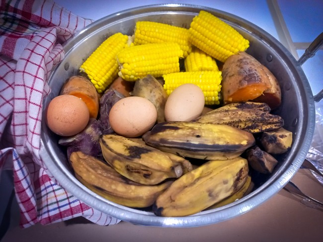 A pot of boiled food contains several ears of corn on the cob, sweet potatoes, bananas, and eggs.