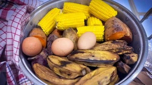 A pot of boiled food contains several ears of corn on the cob, sweet potatoes, bananas, and eggs.