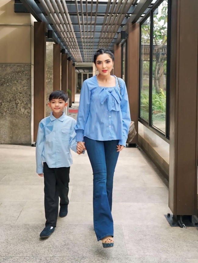 Ashanty memilih blus biru muda berdetail pita besar di bagian dada, memberi kesan lembut dan elegan. Potongan bahunya yang sedikit terbuka membuat tampilannya feminin namun tetap sopan. Ia memadukannya dengan celana jeans lurus berwarna senada, menciptakan tampilan kasual yang tetap chic. Foto: Instagram/@ashanty_ash