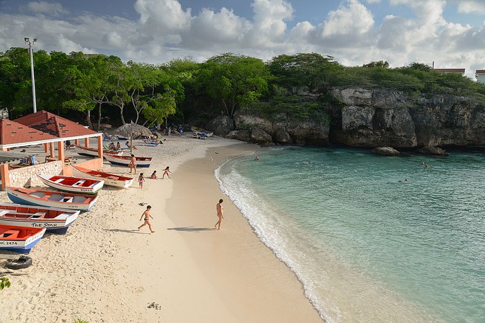 [UNVERIFIED CONTENT] View from the top of the cliffs at Lagun Beach towards the  beach, Curacao, Netherland Antilles.

A couple of persons can be seen walking towards the Caribbeans pristine and turquoise water. Also, it can be seen some colorful fisherman boats on the left side of picture.

The rocky cliffs and a partially cloudy sky can be seen at the background.