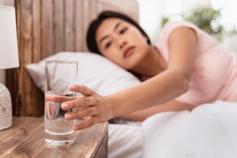 Ilustrasi haus atau minum air saat bangun tidur Healthy Liquid. Asian Lady Drinking Water From Glass Starting Day With Hydration Lying In Bed At Home. Selective Focus
