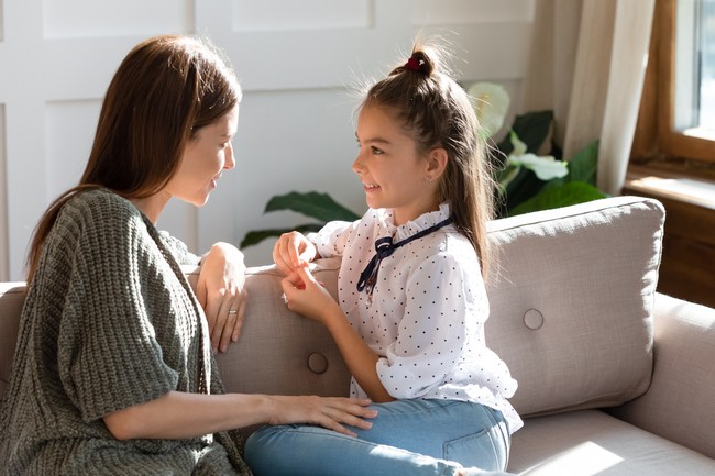 Young Caucasian mom sit on couch with small daughter talk share secrets gossip at home. Happy little girl child speak with mother or nanny enjoy tender intimate moment together. Bonding concept.