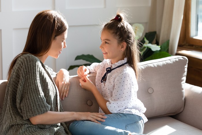 Young Caucasian mom sit on couch with small daughter talk share secrets gossip at home. Happy little girl child speak with mother or nanny enjoy tender intimate moment together. Bonding concept.