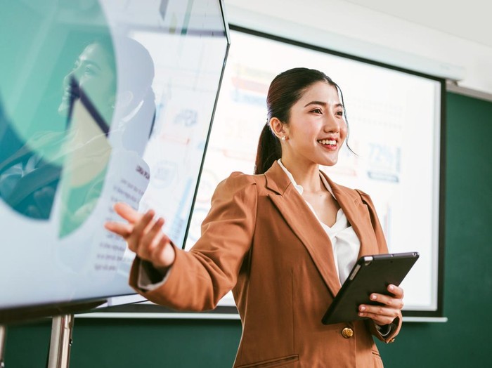 A professional Asian business woman presents data on screens in a board meeting room, engaging her audience with confidence and clarity. A businessman wearing a blazer while using a tablet for meeting
