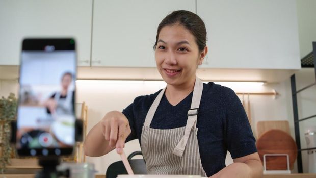 Ilustrasi content creator Cheerful Asian cook mixes food and smiles while creating recipe video content with smartphone for social media followers.