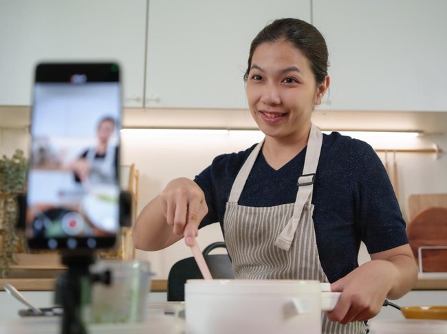 Cheerful Asian cook mixes food and smiles while creating recipe video content with smartphone for social media followers.