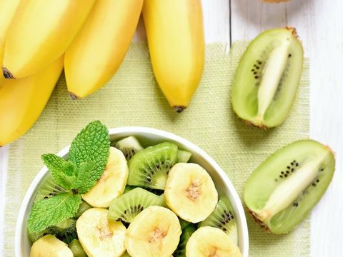 Kiwi and banana salad in bowl and fruits on white wooden table, top view