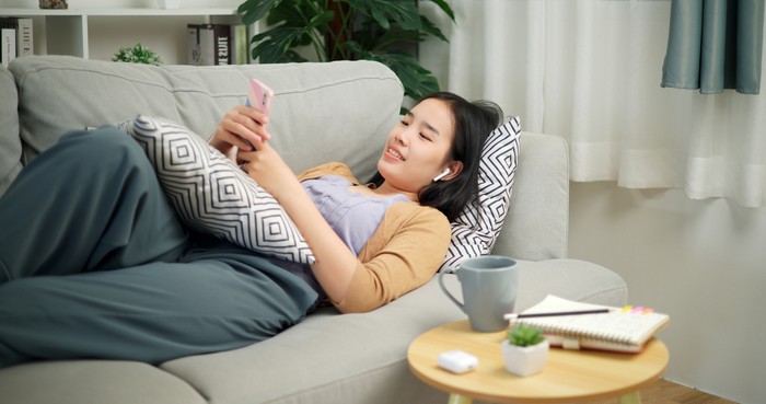 Portrait of Relaxed young Asian woman wear wireless headphones listen to music and sing while lying playing mobile phone on the sofa in the living room at morning ,Free time