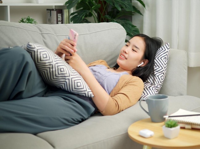 Portrait of Relaxed young Asian woman wear wireless headphones listen to music and sing while lying playing mobile phone on the sofa in the living room at morning ,Free time