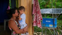 Seorang ibu menggendong anaknya di depan rumah yang kini mendapat listrik dari panel surya, menggantikan ketergantungan lama pada generator diesel yang mahal dan tidak stabil. Lebih dari 990.000 warga Amazon sebelumnya hidup tanpa listrik layak. REUTERS/DIEGO HERCULANO