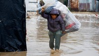 Seorang anak kecil memanggul karung berisi barang miliknya sambil berjalan melewati air. Kondisi ini menggambarkan bagaimana banjir memaksa keluarga memindahkan barang-barang mereka untuk menyelamatkannya dari kerusakan. REUTERS/Mahmoud Issa