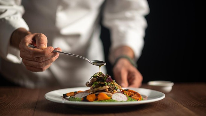 A male chef pouring sauce from the spoon on meal on a black background.