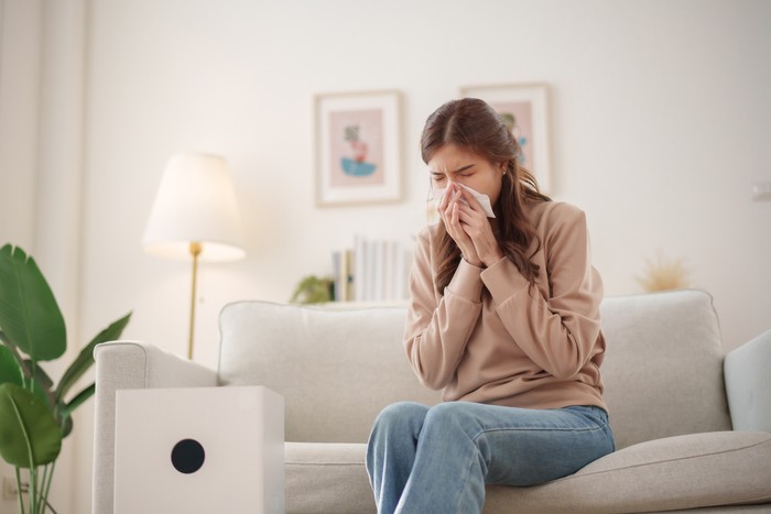 Young Asian woman sits on a couch, sneezing and holding a tissue to her nose while an air purifier operates nearby to improve air quality.