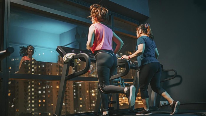low angle rear view body positive woman running treadmill together wiit her female friend in gym at night facing city street light