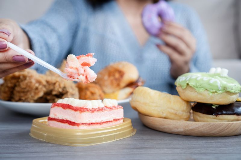 Ilustrasi makan junk food dan manis Woman eating too much fast food like burgers, fried chicken, donuts, and sweets, showing signs of binge eating and emotional stress with unhealthy food habits