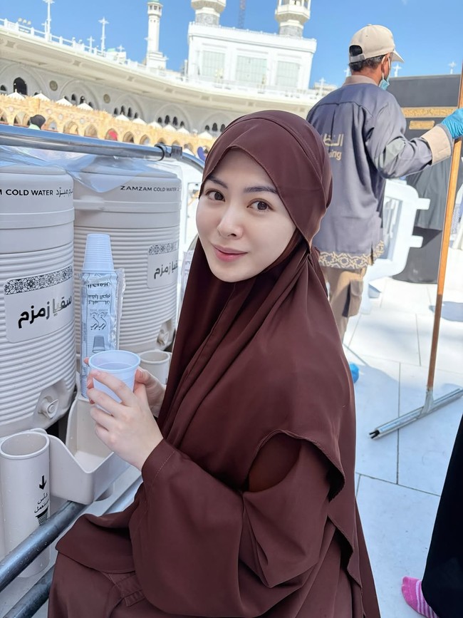 Ayana tampil tanpa makeup dengan hijab instan berwarna cokelat tua yang memiliki potongan praktis dan menutupi dada. Gaya ini memberikan kesan nyaman dan tetap rapi saat ia sedang beristirahat sambil menikmati air zam-zam di area Masjidil Haram. Foto: Dok. Instagram @xolovelyayana.