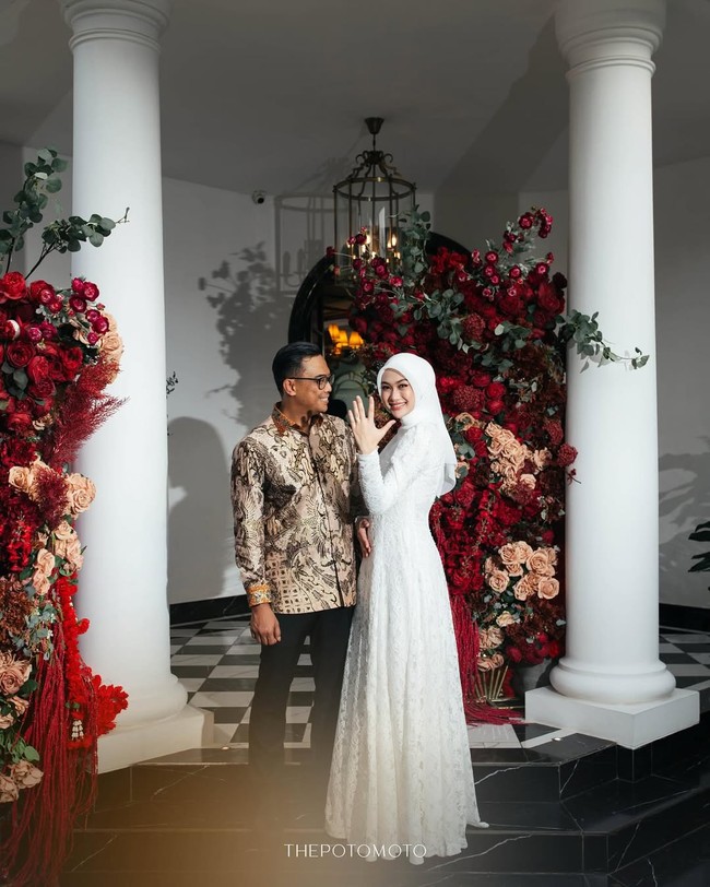 Indah Nada Puspita berpose anggun sambil menunjukkan cincin pertunangan di jari manisnya dengan latar belakang dekorasi bunga bernuansa merah yang elegan. Senyum bahagia terpancar jelas dari wajahnya saat bersanding dengan Dhani Yahya yang tampil serasi mengenakan kemeja batik motif cokelat. Foto: Dok. Instagram @indahnadapuspita.
