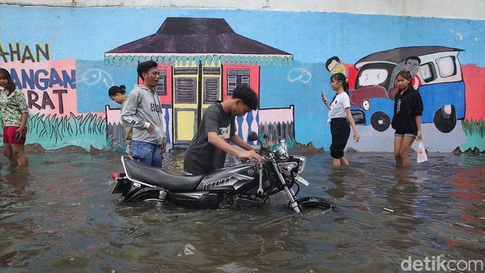 Pengendara mendorong sepeda motor mogok saat melintasi genangan banjir di kawasan Mangga Dua, Jakarta Utara, Senin (12/1/2026). Tingginya air membuat mesin kendaraan mati.