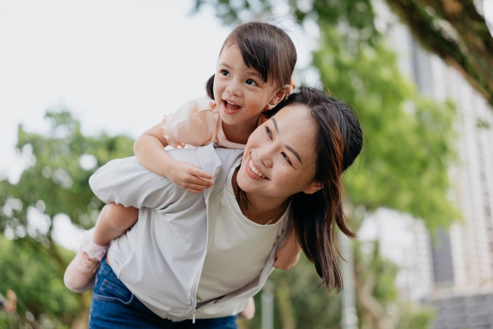 An Asian Chinese mother playing piggyback riding with her young daughter in a public park