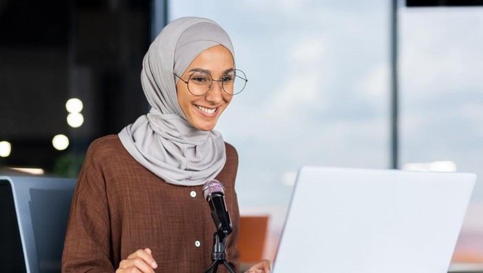 A young Muslim woman in a hijab is sitting in an office in front of a laptop and a microphone. records a podcast, a blog, talks on a video call, gives an interview, goes through an online interview.