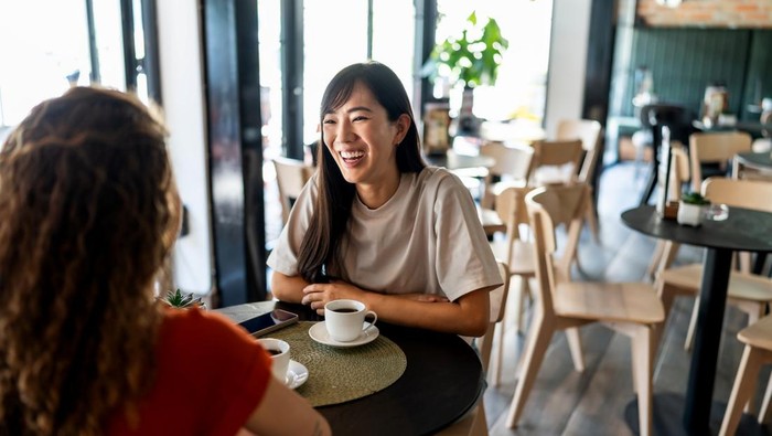 Two young women sharing laughter and engaging in a lively conversation while sipping coffee together at a bright, modern cafe