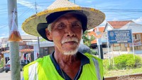 Ia mengenakan caping dan rompi hijau untuk melindungi diri dari panas terik. Tak ada informasi resmi jadwal kereta api yang akan melintas.