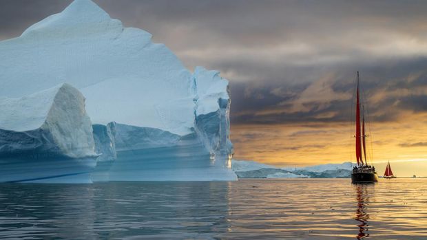 Perahu layar merah berlayar di antara gunung es terapung di gletser Teluk Disko, Ilulissat, Greenland, dengan matahari terbenam oranye selama musim panas kutub tengah malam.