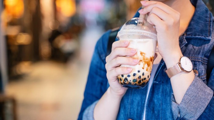 Young girl in denim jacket is drinking brown sugar flavored tapioca pearl bubble milk tea with glass straw in night market of Taiwan, close up, bokeh