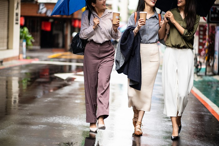 Three female friends walking on the street, drinking coffee on the move, talking and smiling lively on rainy day