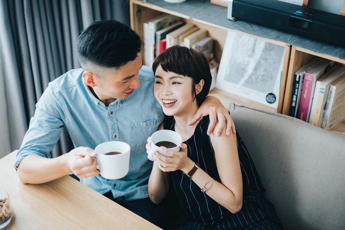 Loving young Asian couple chatting and having coffee while relaxing in living room at home