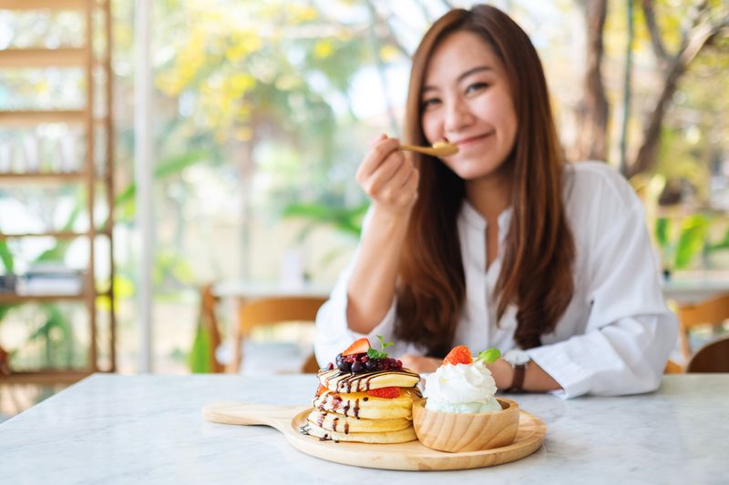 Ilustrasi penyuka makanan manis A beautiful asian woman eating ice cream and a mixed berries pancakes with whipped cream by wooden spoon