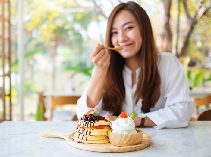 A beautiful asian woman eating ice cream and a mixed berries pancakes with whipped cream by wooden spoon