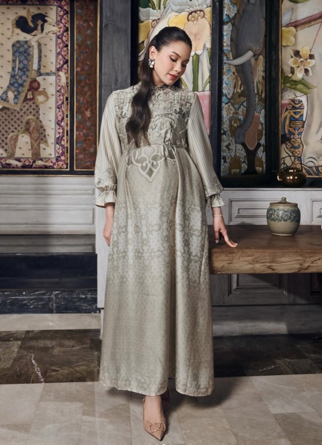 Alyssa tampil dengan gamis panjang berpotongan A-line yang jatuh flowy hingga mata kaki, memberi kesan anggun dan lembut. Didominasi warna sage green keabu-abuan, gamis tersebut memiliki motif etnik bernuansa floral yang cantik. Bagian perutnya tampak dibuat lebih lebar untuk menyesuaikan babyy bump Alyssa yang sudah semakin besar. Foto: Instagram/@alyssadaguise/@geulis.id