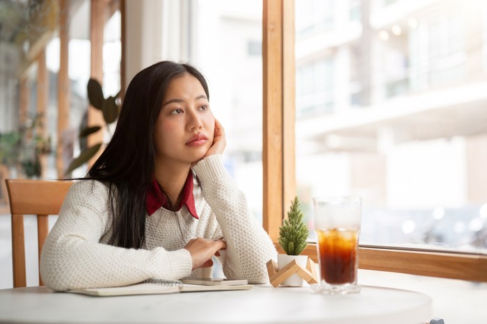 An asian woman is putting her hand under the chin, looking bored or unhappy while waiting for someone at the round marble table. Study or Working in the cafe.