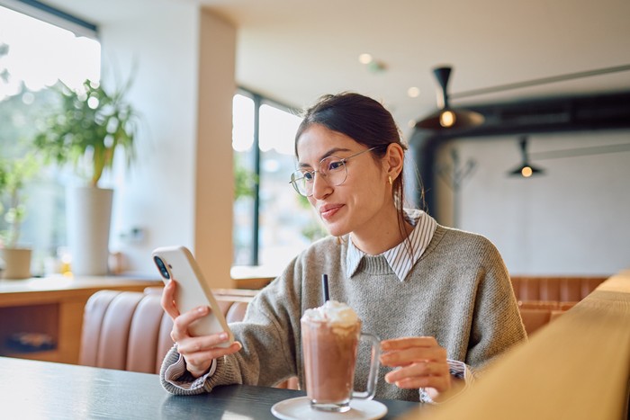 Young woman relaxing at a cafe, holding a smartphone, interacting with digital content while sipping a hot chocolate