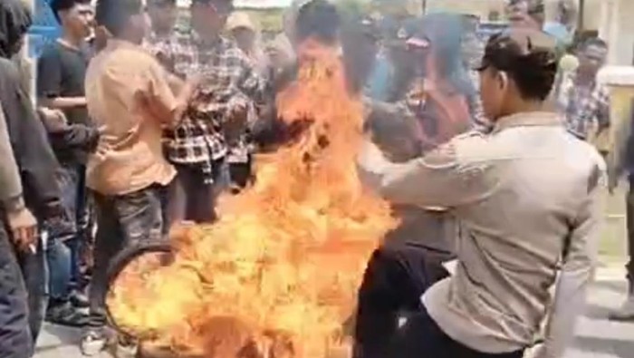 Seorang anggota polisi menendang ban bekas yang terbakar saat demo di Kabupaten Muna Barat, Sulawesi Tenggara (Sultra). Ban yang terbakar mengarah ke massa demo hingga lima orang dilaporkan mengalami luka bakar.
