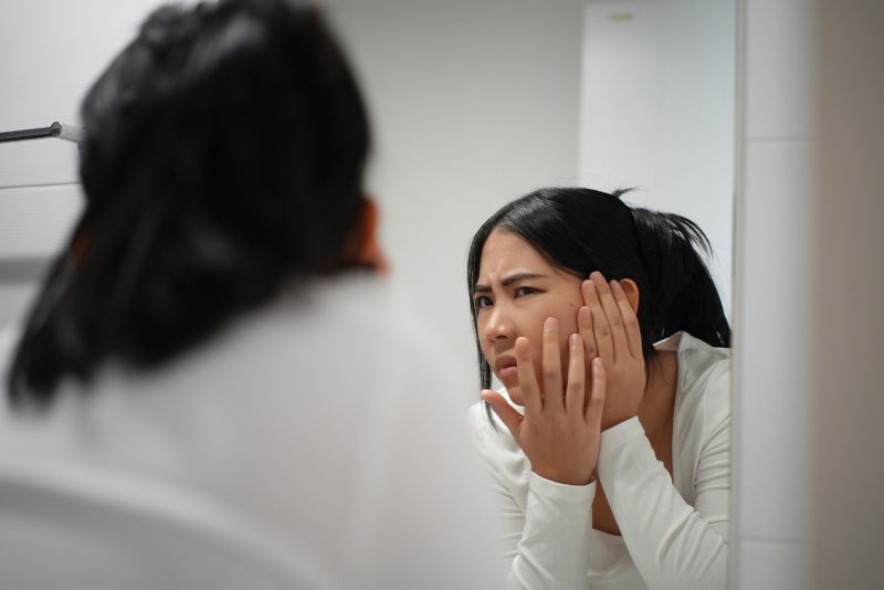 Unhappy young woman worried her wrinkle acnes on face and eyes bag while standing in front the bathroom mirror, female checking reflection her face and disappointed with aging face problems.