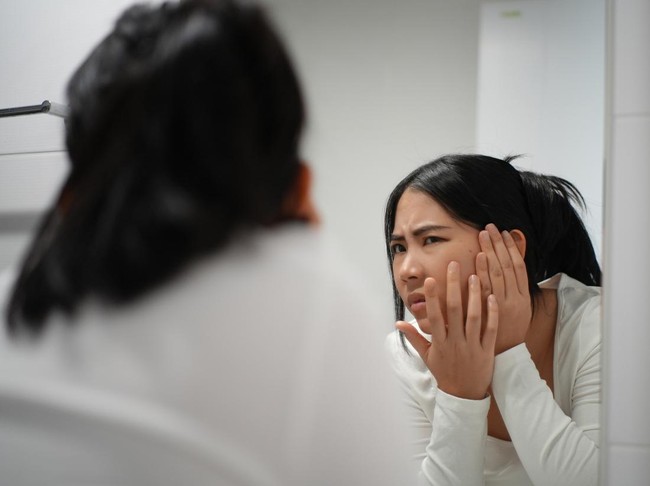 Unhappy young woman worried her wrinkle acnes on face and eyes bag while standing in front the bathroom mirror, female checking reflection her face and disappointed with aging face problems.