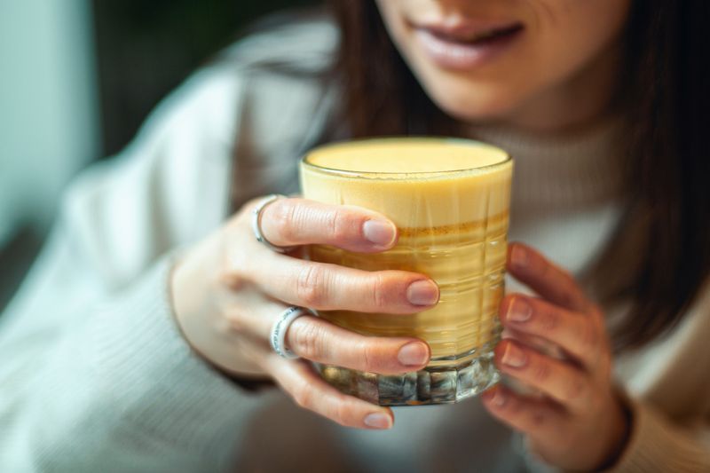 kunyit Young caucasian woman in warm sweater is smiling and holding a glass of hot turmeric milk on table of coffee shop. Enjoyment of drinking hot beverage near window on weekend. Copy space
