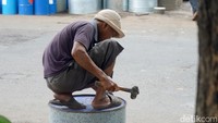 Seorang pekerja sedang memotong drum dan membentuk logam secara manual. Palu, pahat, dan kikir digunakan bergantian, dengan irama pukulan yang konstan mengikuti bentuk yang diinginkan.