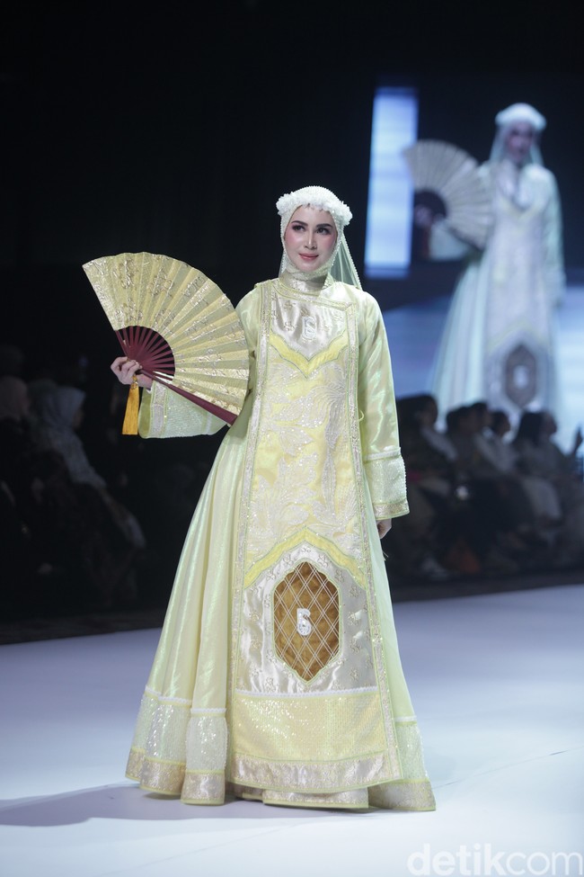 Arumi Bachsin tampil dalam balutan busana muslim bernuansa kuning keemasan karya Lina Sukijo, memadukan keanggunan gaya modern dengan sentuhan kipas tradisional yang mewah. Dengan senyum yang khas, Arumi membawakan koleksi tersebut di atas panggung runway dengan penuh percaya diri, menonjolkan detail bordir yang rumit dan siluet busana yang megah. Foto: Gilang Faturahman/detikFoto.
