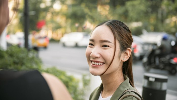 Young asian woman smiling and looking at her friend while having a conversation on a city street, with blurred cars and trees in the background