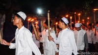 Anak-anak hingga orang dewasa berjalan beriringan sambil membawa obor, sebagian mengenakan busana muslim, peci, dan kerudung.